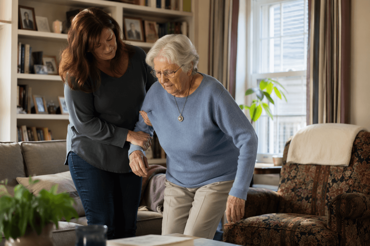 An older woman being gently assisted to stand from a sofa by a home care aide in a warm living room