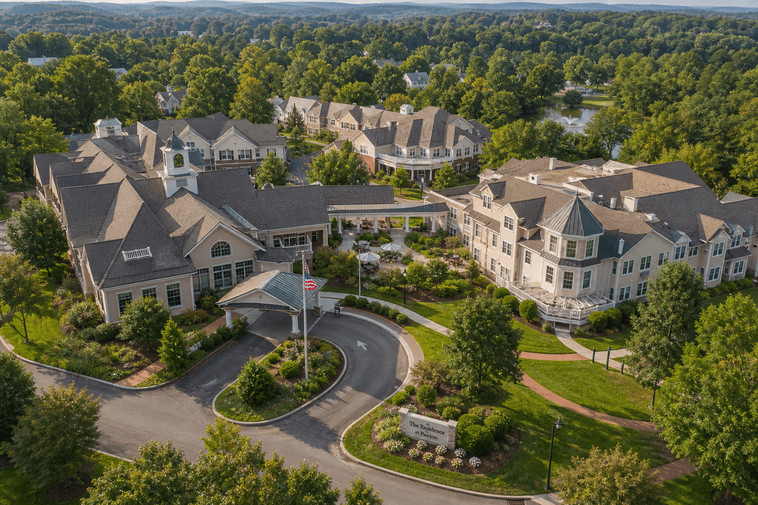 Elevated view of the D'Youville Life & Wellness Community campus in Lowell, Massachusetts, showing multiple connected senior care buildings surrounded by mature trees, landscaped grounds, and walking paths under warm daylight, with a residential community atmosphere