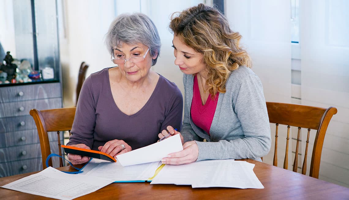 An adult daughter sits beside her elderly mother at a wooden table, both looking at financial paperwork and a notebook together.