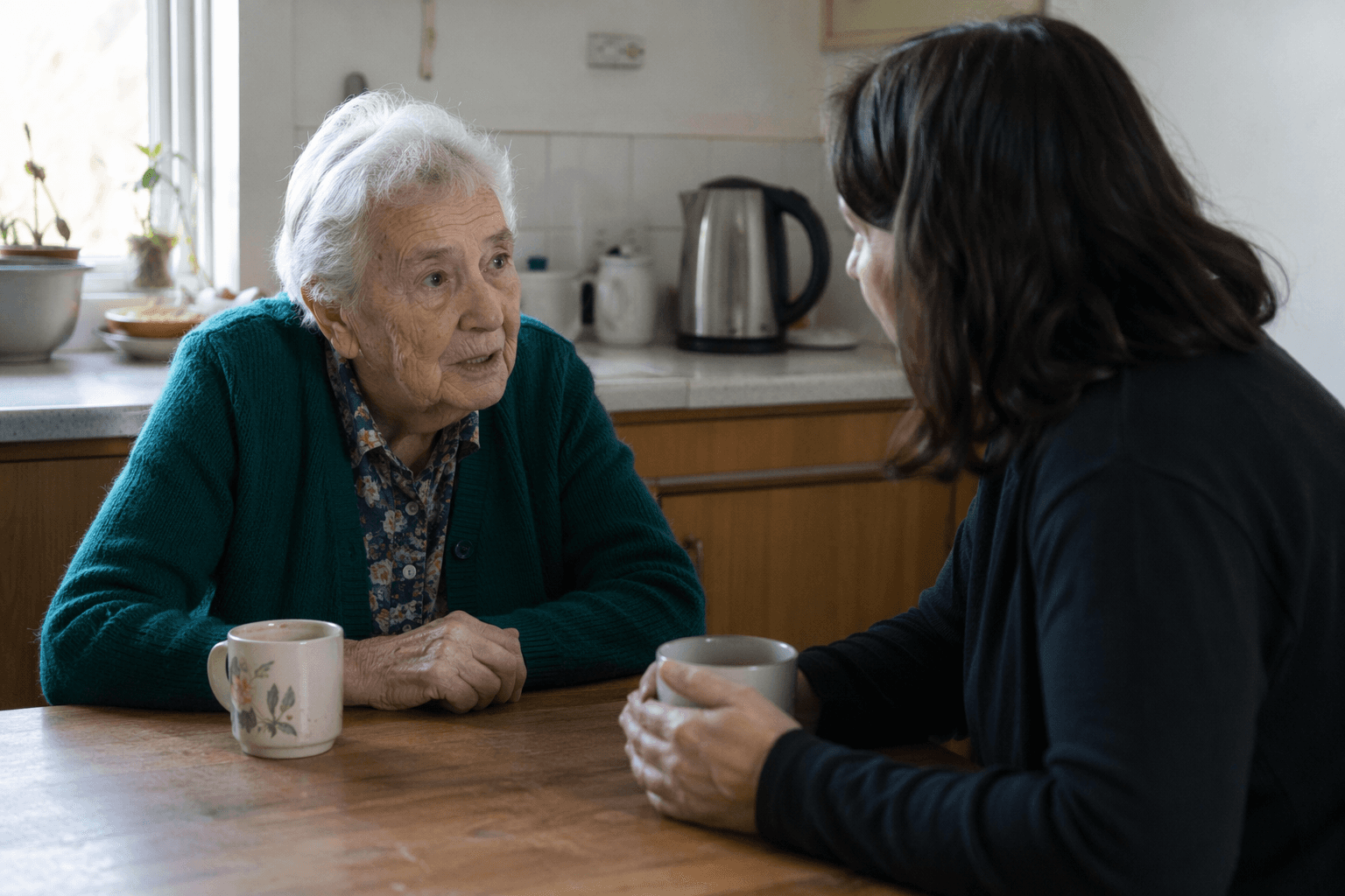 An older woman and younger companion talking at a kitchen table with mugs nearby