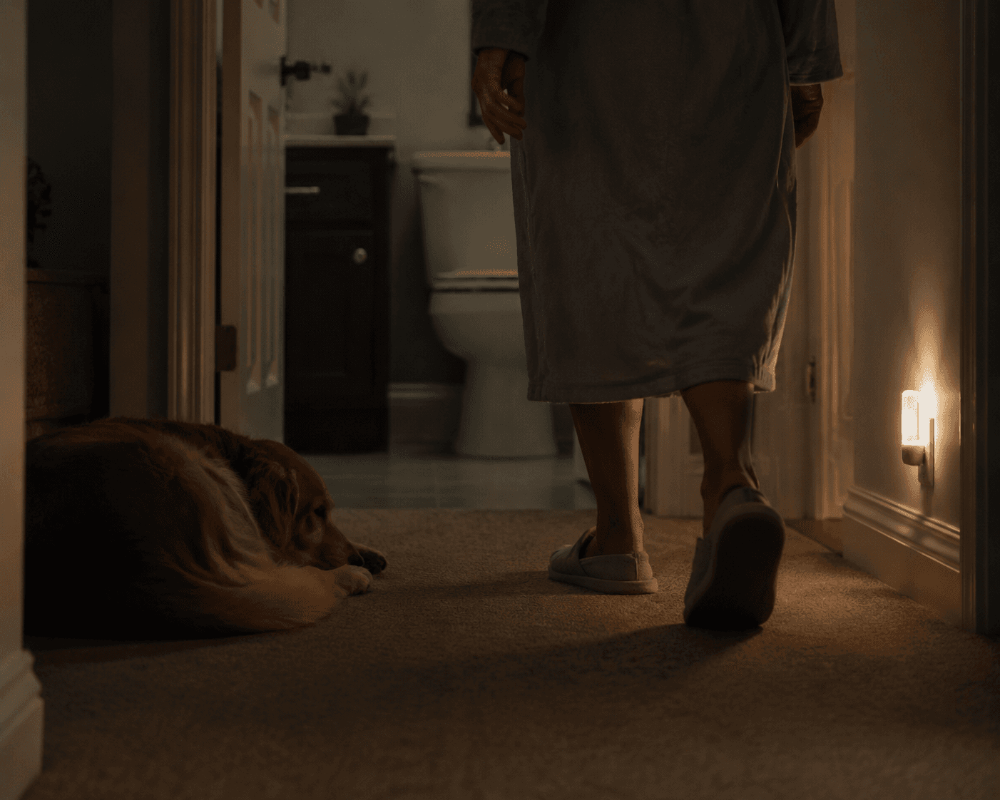 Dim hallway at night with an older adult walking toward a bathroom while a dog lies in the shadowed path