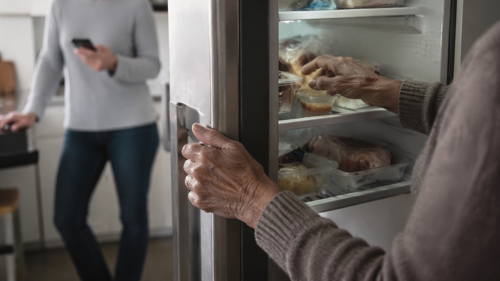 Older adult opening a refrigerator while a caregiver checks a smartphone nearby