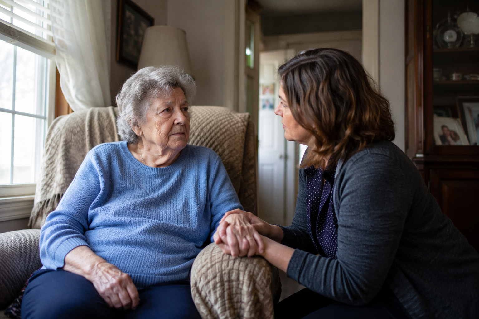 An elderly woman sitting beside a middle-aged caregiver in a modest living room, holding hands in a quietly worried scene