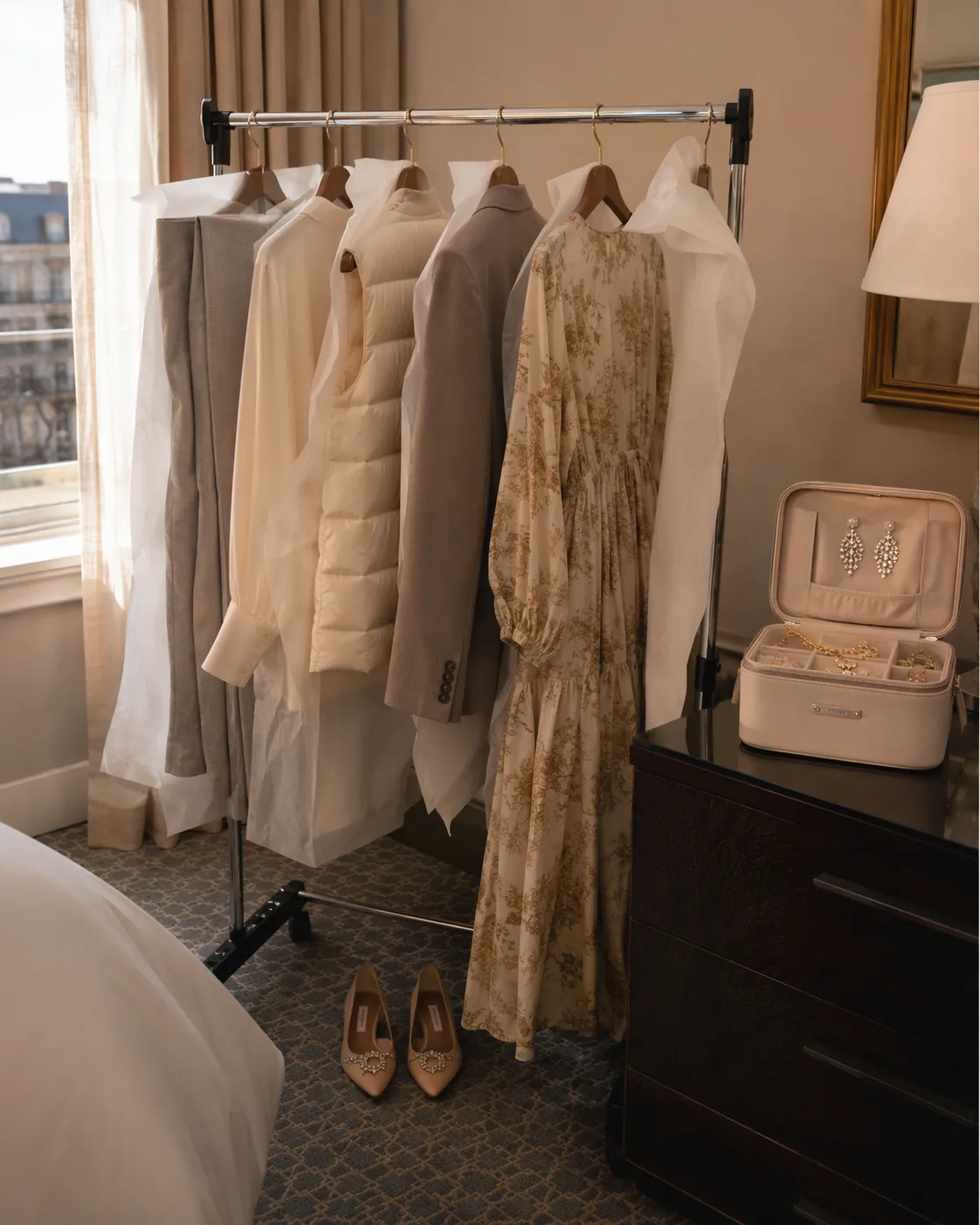 A hotel room garment rack holds planned outfits with tissue paper, jeweled flats, and a small jewelry case nearby