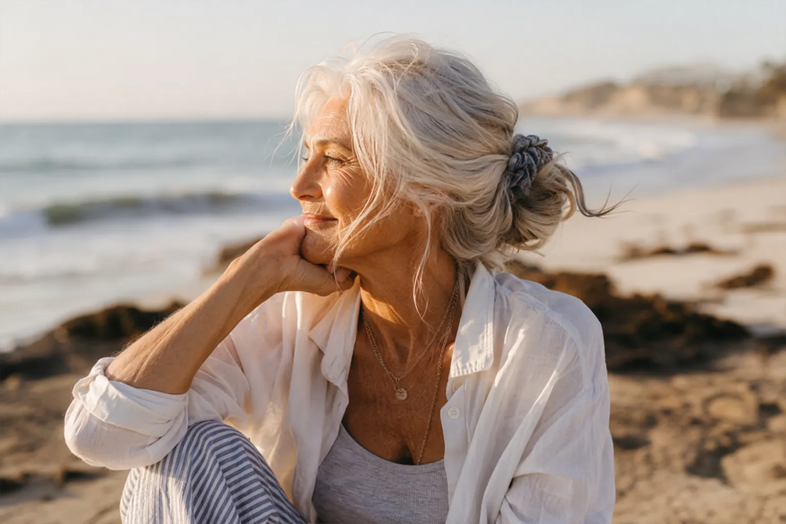 Older woman with fine silver-white hair in a loose low bun secured with a fabric scrunchie on a sunny beach