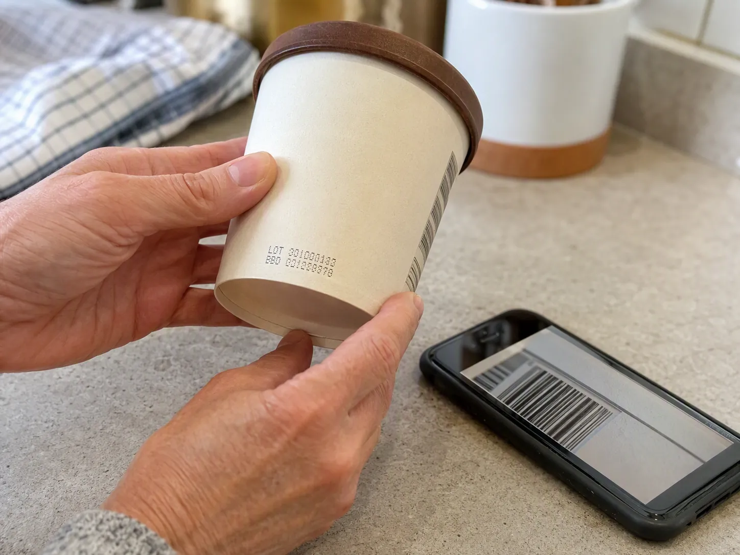 Hands turning an ice cream carton to read small lot and date codes while a phone shows a barcode