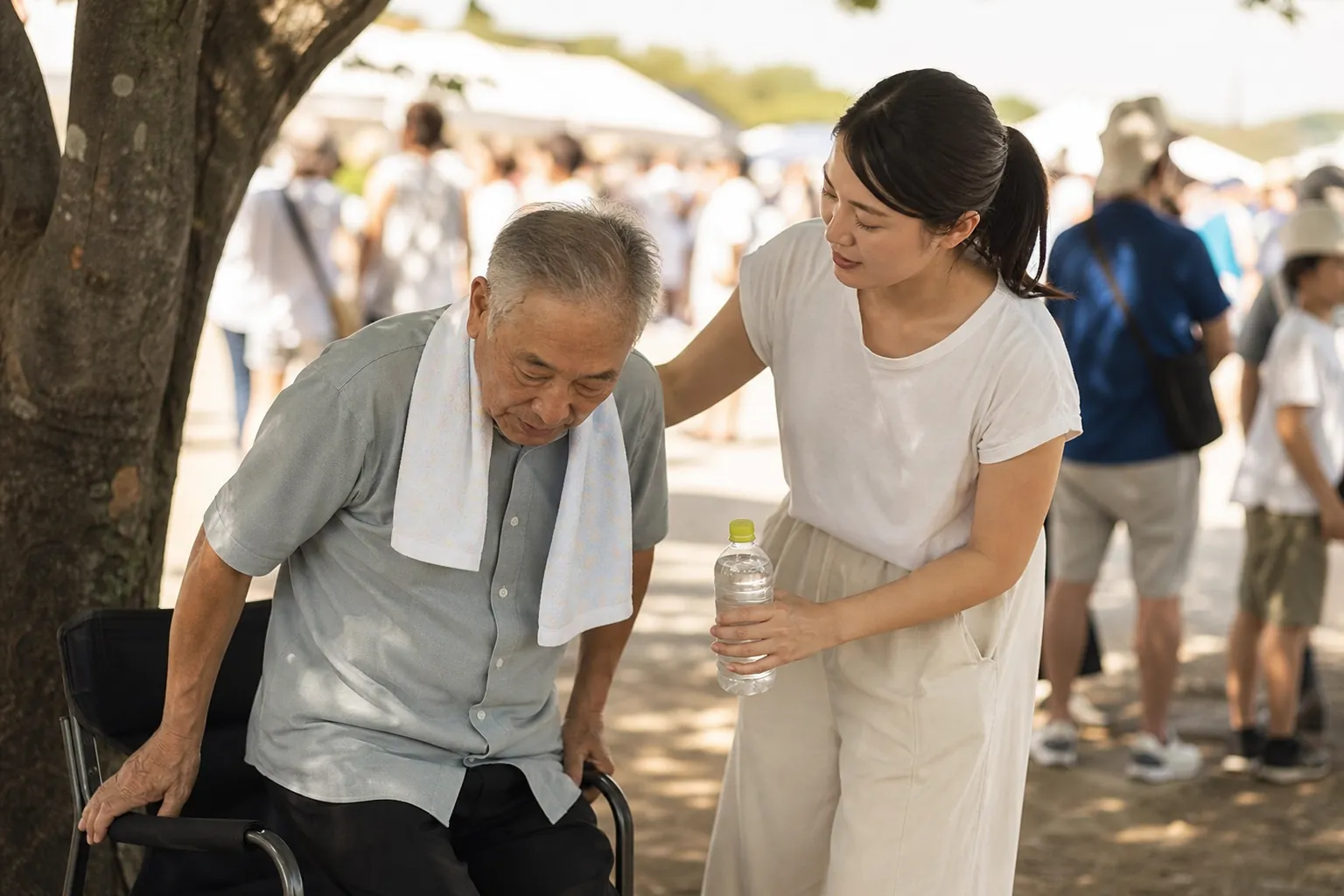 Adult daughter helping her older father sit in the shade at an outdoor event with a cooling towel and water bottle