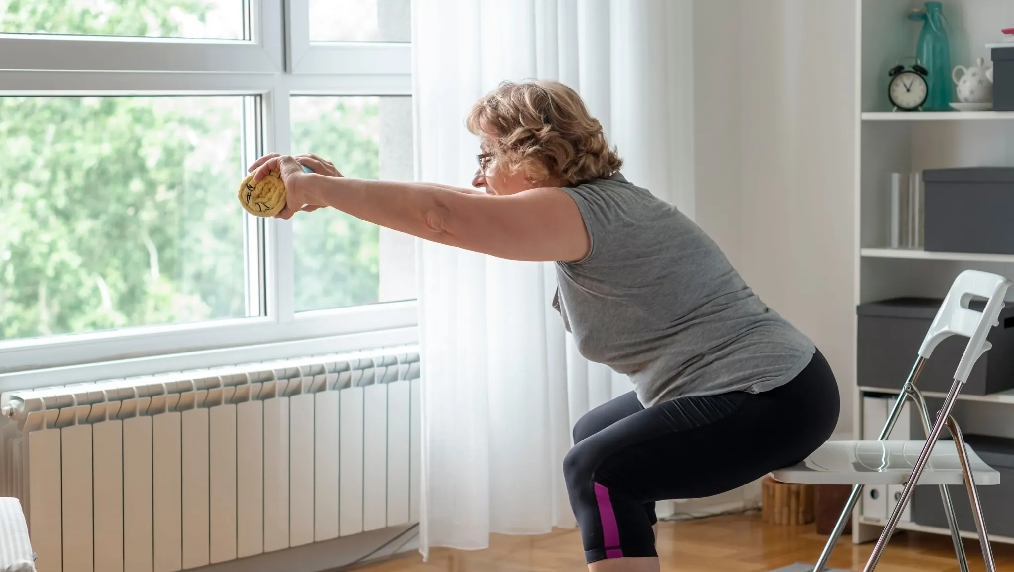 Two older women performing seated strength exercises on chairs in a bright home living room