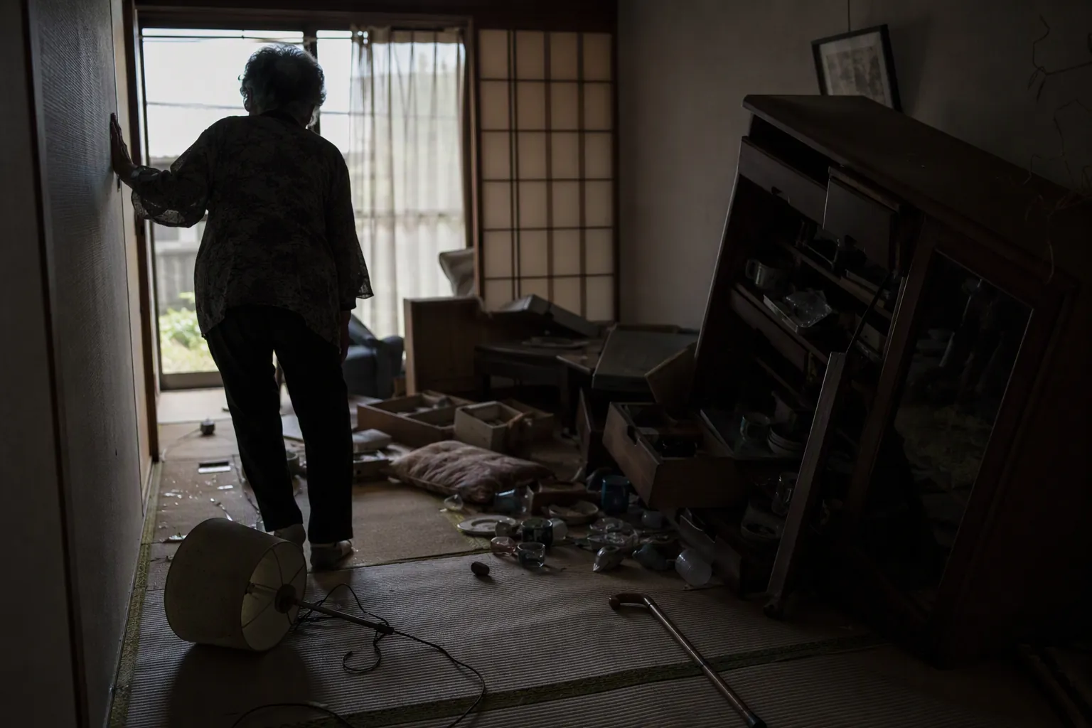 Dim Japanese room after an earthquake with fallen lamp, tilted cabinet, cluttered tatami floor, cane, and an older person steadying against the wall