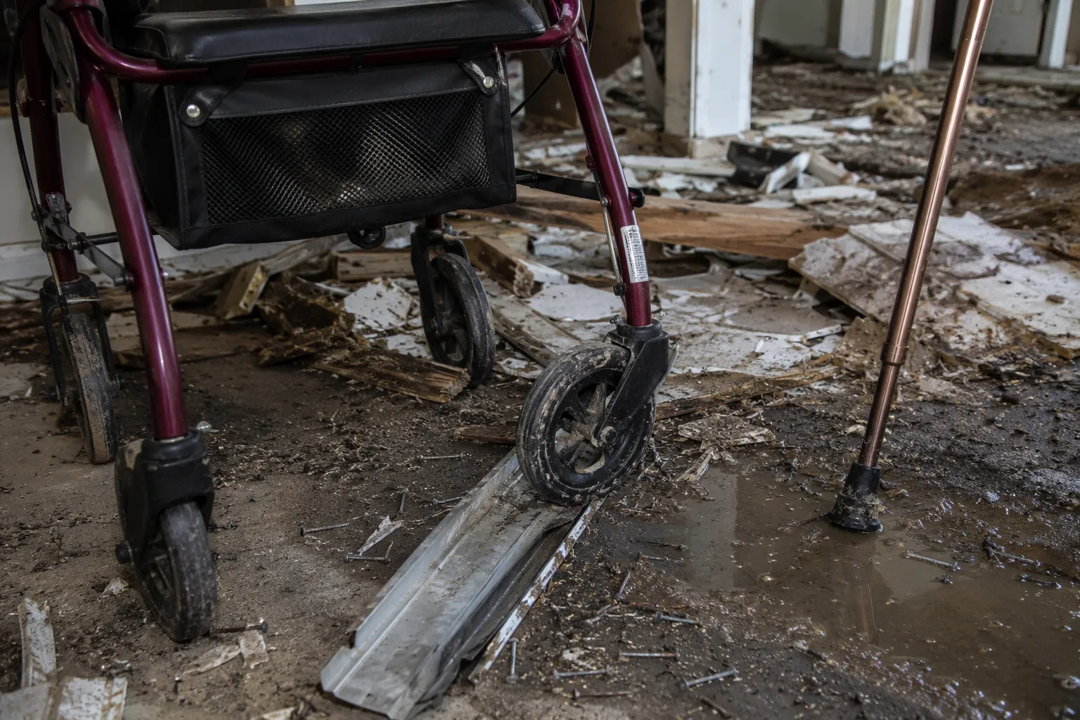 Rollator walker and cane on a tornado-damaged floor with nails, wet mud, splintered wood, and roofing grit