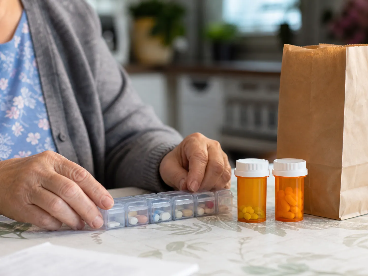 Older person's hands arranging a weekly pill organizer beside prescription bottles and an empty pharmacy bag