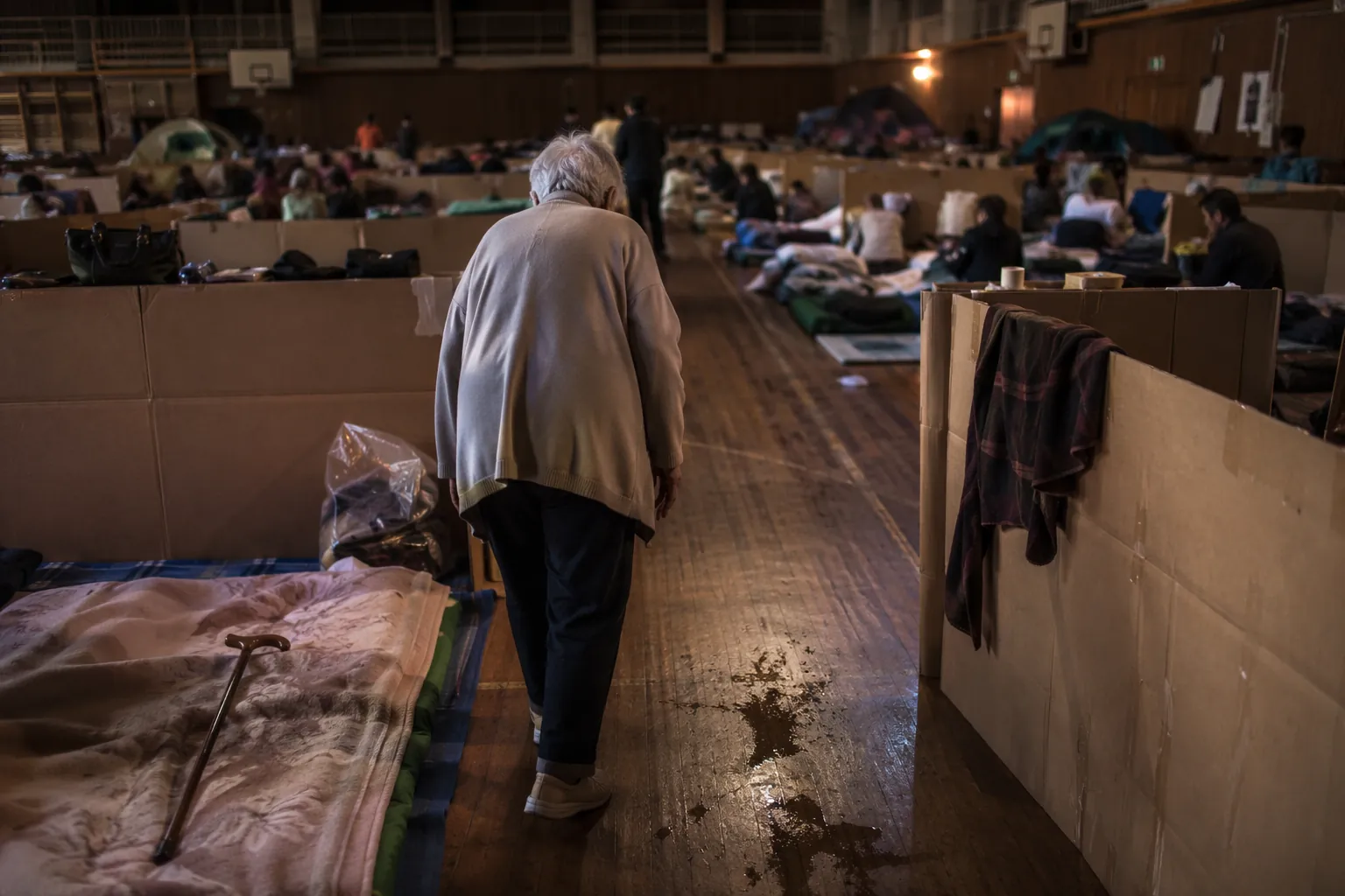 Older adult navigating a dim crowded shelter gymnasium with narrow aisles, a cane nearby, scattered debris, and a wet patch on the floor