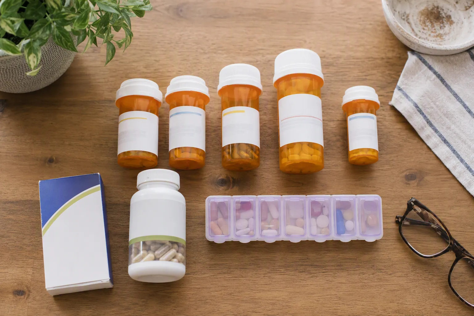 Prescription bottles, over-the-counter medicine, supplement jar, weekly pill organizer, and reading glasses on a kitchen table