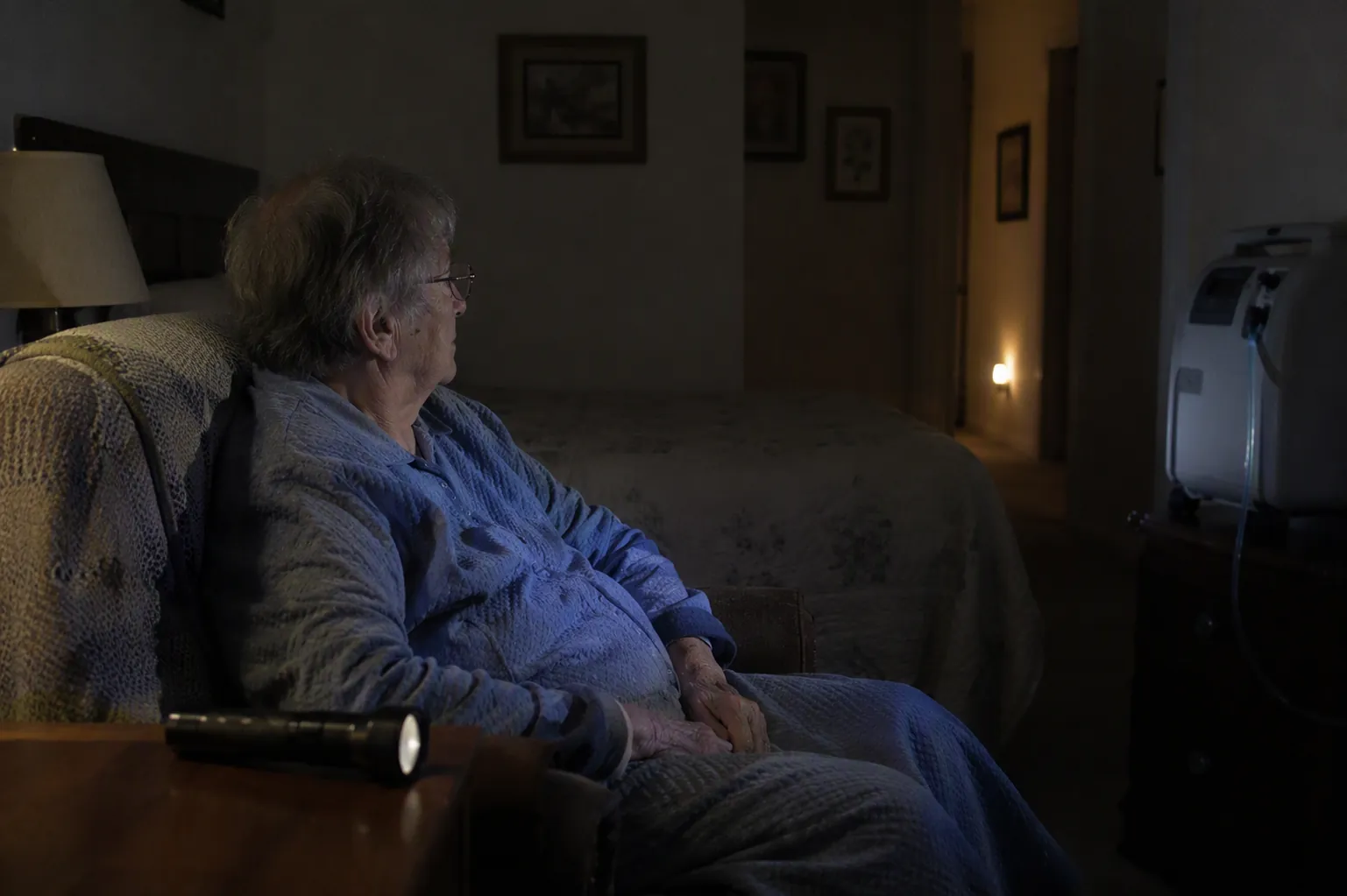 Elderly person seated near a bed during a power outage with a flashlight within reach and medical equipment nearby