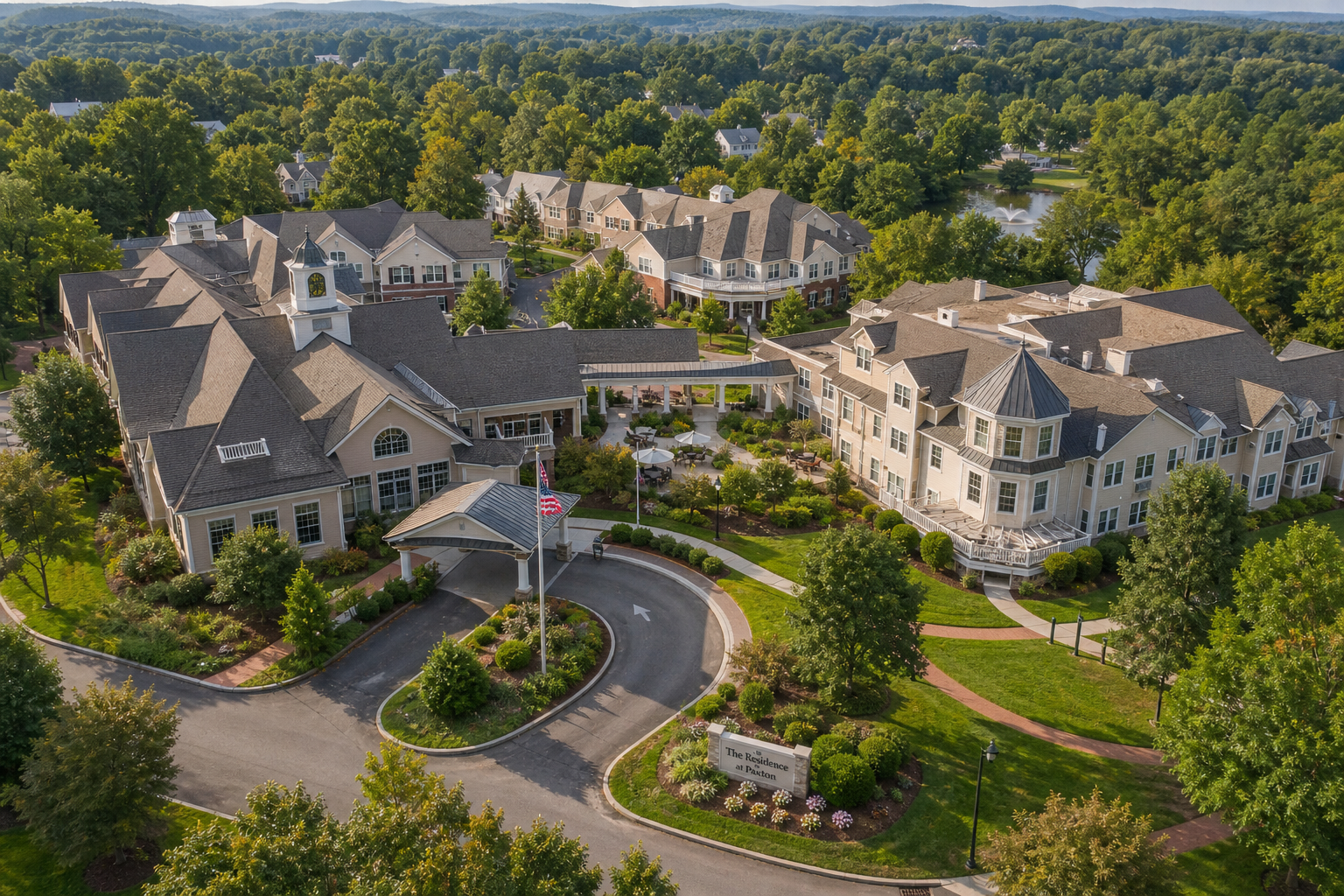 Elevated view of the D'Youville Life & Wellness Community campus in Lowell, Massachusetts, showing multiple connected senior care buildings surrounded by mature trees, landscaped grounds, and walking paths under warm daylight, with a residential community atmosphere