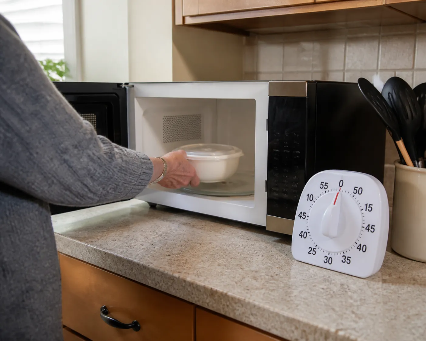 Older adult placing a covered microwave-safe bowl into a microwave with a large-print timer nearby and a clear kitchen floor