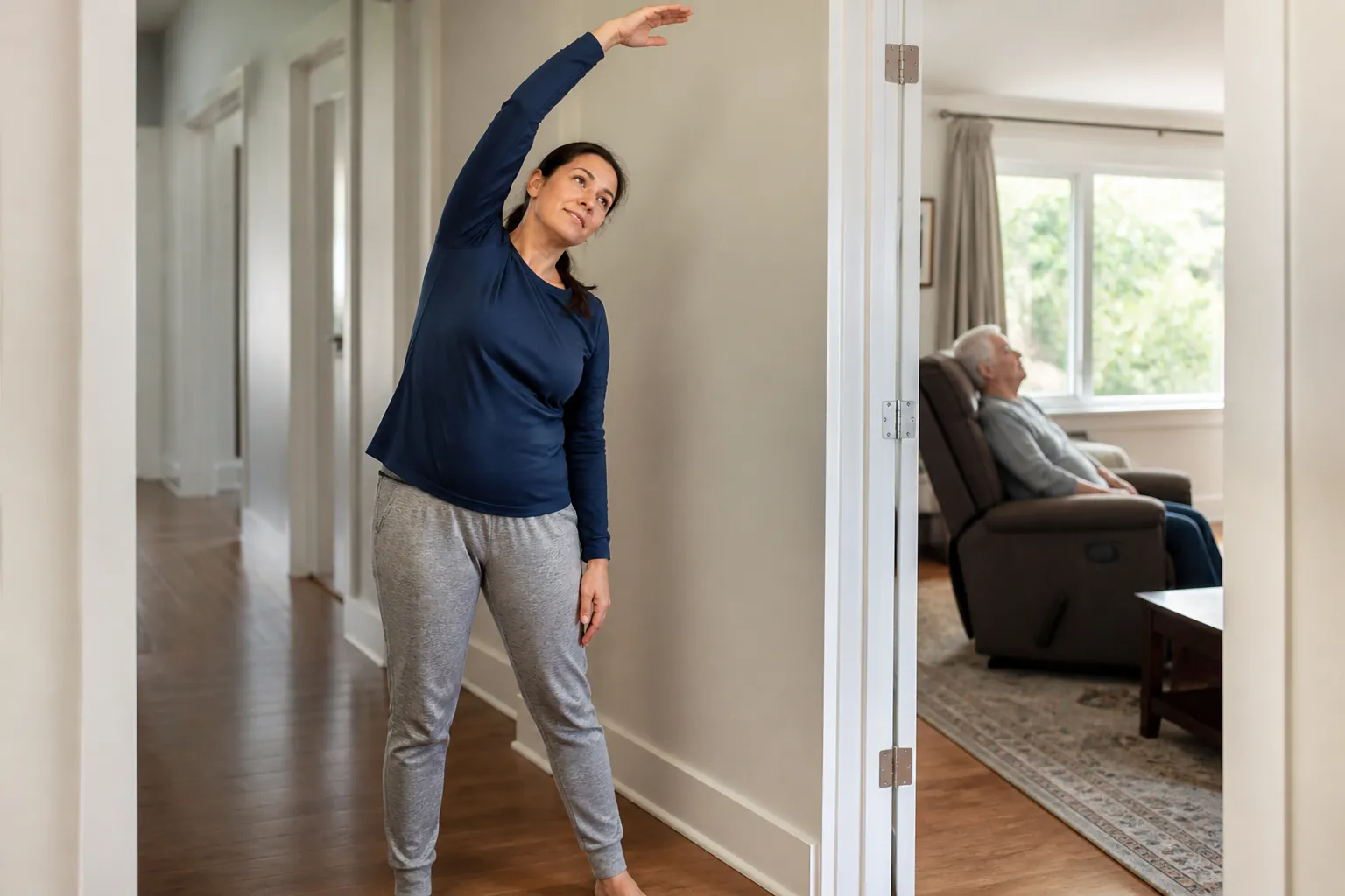 A caregiver stretches gently in a hallway while an older adult rests in a recliner within sight.