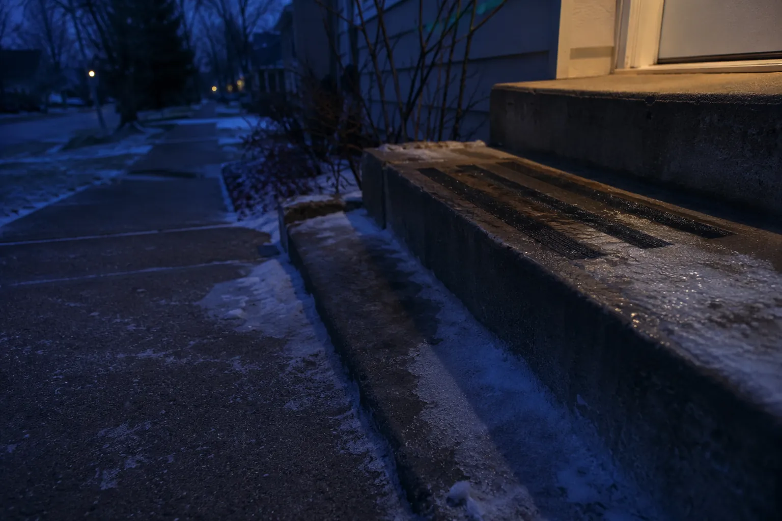 A dark winter step and walkway with ice and traction strips, while dim porch light fails to reach the bottom stair