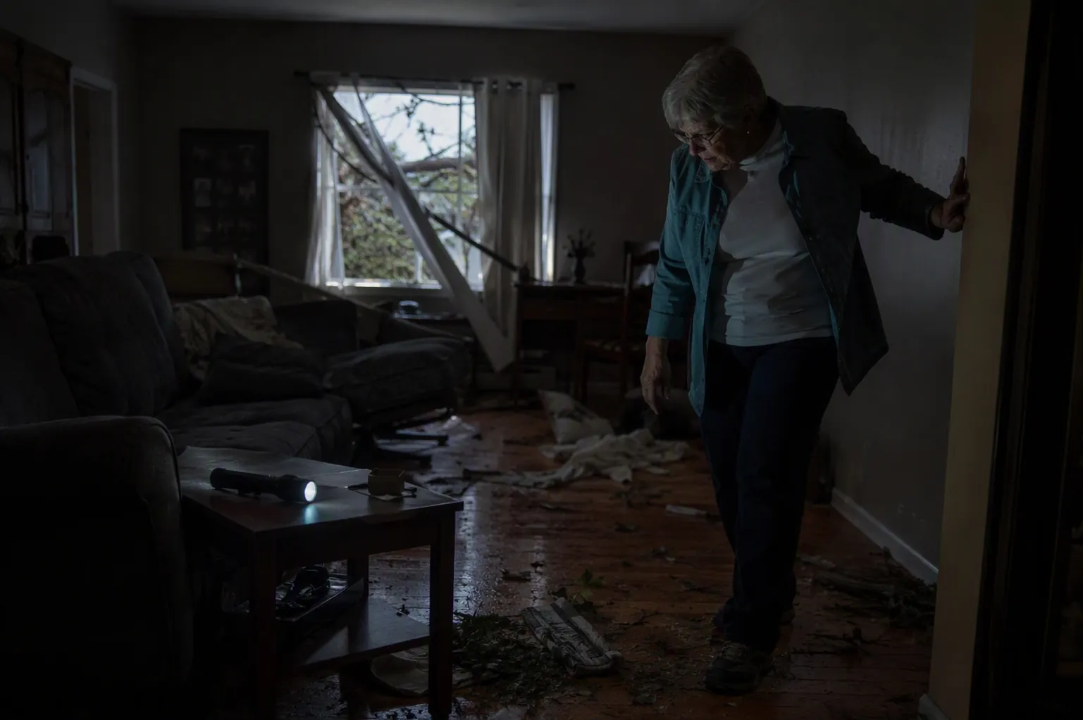Older adult standing cautiously in a darkened post-tornado living room with debris, shifted furniture, sturdy shoes, and a flashlight nearby