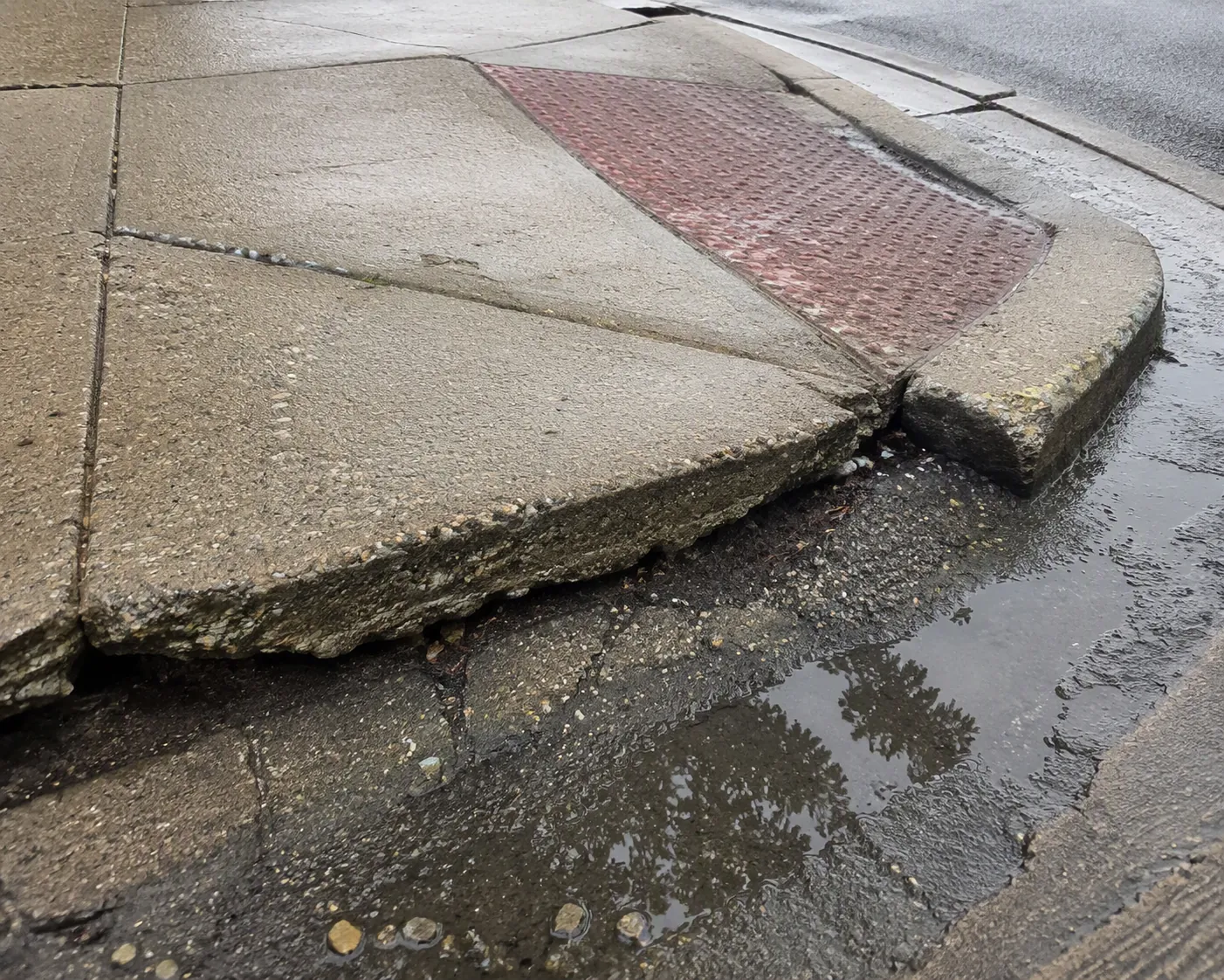 Raised and cracked downtown sidewalk slab with a worn curb ramp and shallow puddle