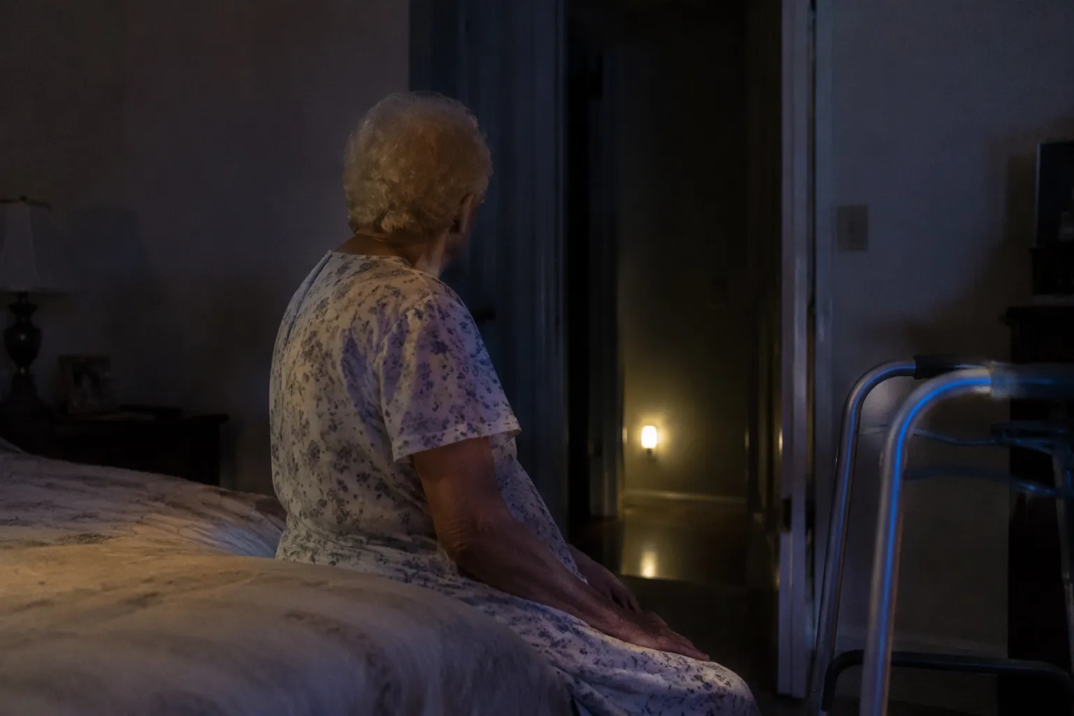 Older adult sitting on the edge of a bed at night, looking toward a dark hallway with a walking aid nearby