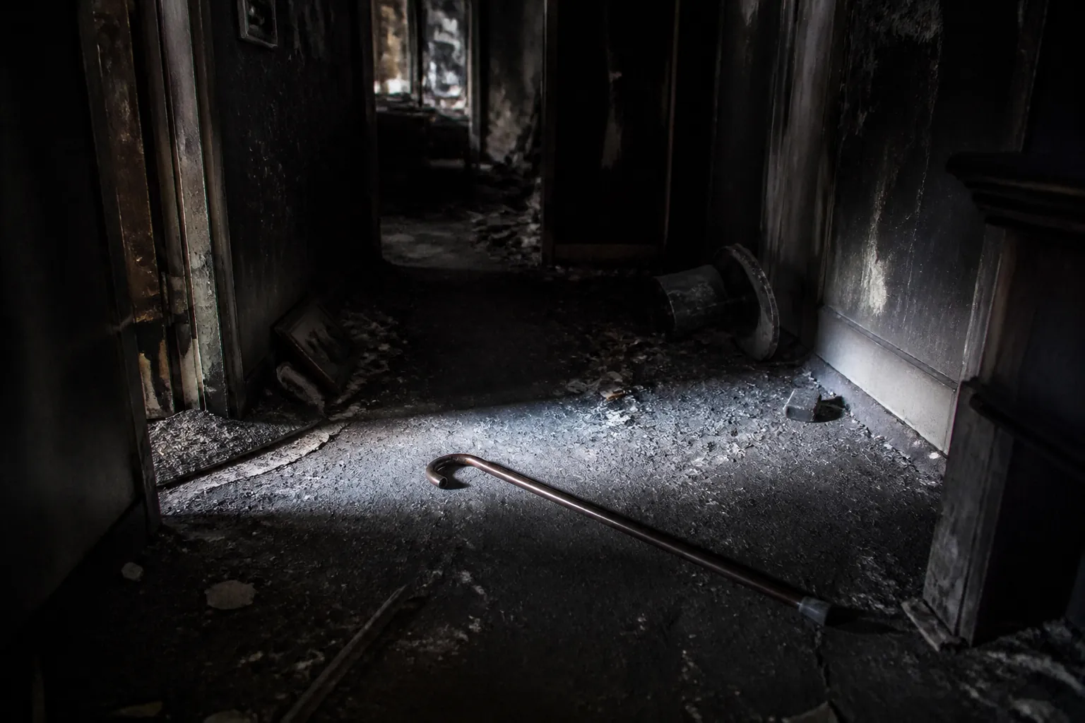 Dark ash-damaged hallway with scattered debris, a cane on the floor, and a flashlight beam showing trip hazards