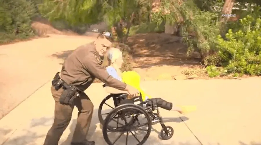 Responders evacuate an elderly wheelchair user as wildfire smoke rises nearby