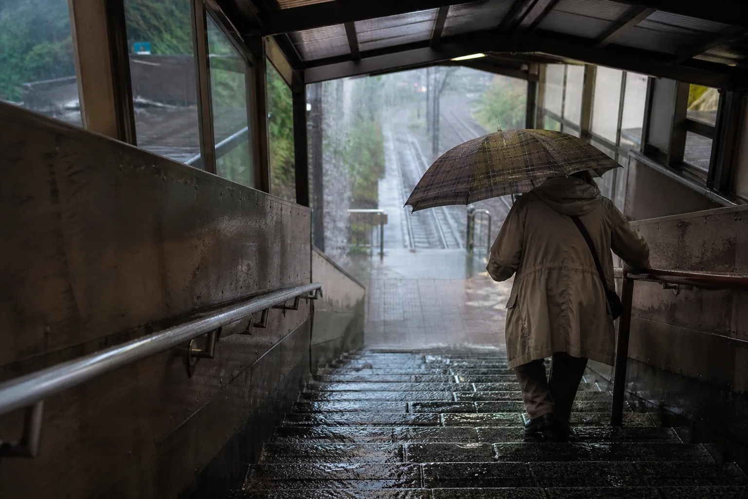 Older traveler gripping a handrail on a wet Japanese train station staircase during heavy rain